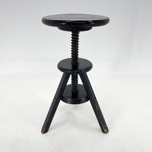 May include: Black wooden adjustable stool with a round seat and three legs. The stool has a screw mechanism to adjust the height.