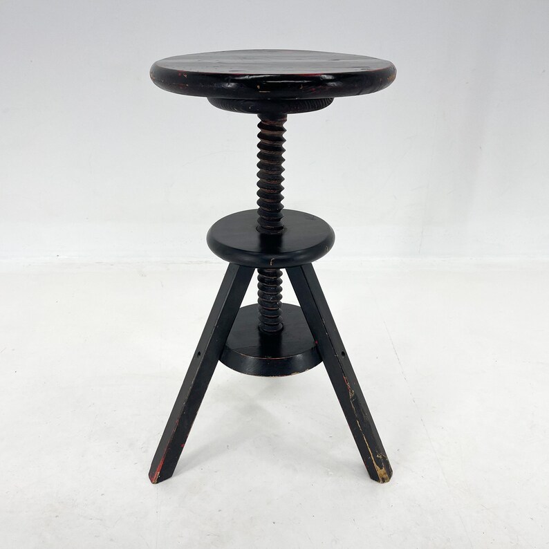 May include: Black wooden adjustable stool with a round seat and three legs. The stool has a screw mechanism to adjust the height.