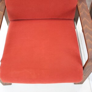 May include: Two mid-century modern chairs with a vibrant red seat and backrest. The wooden frames have a warm, brown finish. The chairs are set against a white background, highlighting their design.