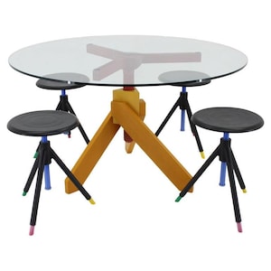 Pode incluir: Um conjunto de jantar moderno com uma mesa redonda de vidro e uma base de madeira. A base tem um design geométrico. Quatro bancos pretos com pernas coloridas rodeiam a mesa. A mesa tem cerca de 120 cm de diâmetro.