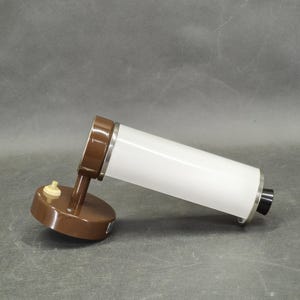 Può includere: Una lampada da parete vintage. La lampada ha una base e un braccio marroni, un paralume cilindrico bianco e un piccolo interruttore on/off. Presenta finiture argentate e un tappo terminale nero. Il design è semplice e funzionale.