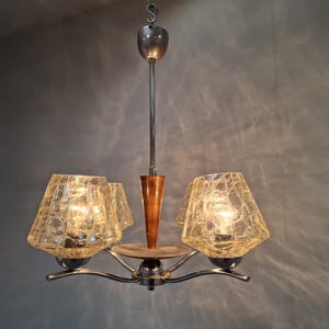 Può includere: Un lampadario vintage a tre luci. L'apparecchio ha una finitura cromata con un accento in legno. I paralumi sono in vetro ambrato strutturato. Il lampadario è sospeso a una catena di metallo.