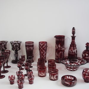 Könnte beinhalten: Eine Sammlung rubinroter Glaswaren, darunter Weingläser, Becher, ein Krug, eine Karaffe und eine Schüssel. Die Glaswaren weisen aufwendige Ätzmuster auf und sind auf einer weißen Oberfläche angeordnet.