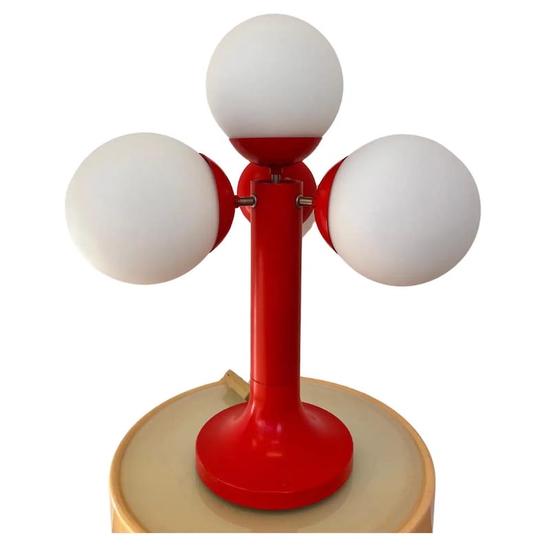 May include: A red table lamp with three white globe shades. The lamp has a central red stem and a round base. The shades are evenly spaced around the stem. The lamp is on a light-colored surface.