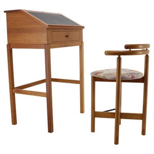 May include: A wooden writing desk with a slanted top and a single drawer, accompanied by a matching chair. The desk features a dark surface on top, and the chair has a floral patterned seat. The set is crafted from light brown wood.