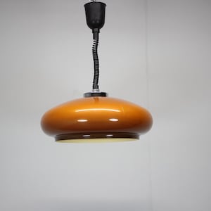 May include: A vintage orange pendant light with a rounded, saucer-like shade. The light fixture has a black, flexible cord and a black ceiling mount. The shade is a glossy, warm orange color, and the bottom rim is a darker brown.