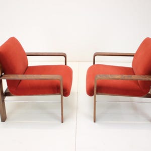 May include: Two mid-century modern armchairs. Each chair features a vibrant red upholstered seat and backrest, complemented by a dark wood frame and armrests. The chairs have a minimalist design with a sleek, curved silhouette and metal supports.