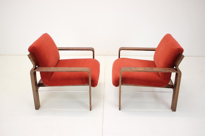 May include: Two mid-century modern armchairs. Each chair features a vibrant red upholstered seat and backrest, complemented by a dark wood frame and armrests. The chairs have a minimalist design with a sleek, curved silhouette and metal supports.