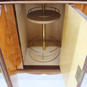 May include: A wooden cabinet with two doors open, revealing a bar area with a rotating shelf and a brass pole. The cabinet has a wood veneer with a geometric pattern.