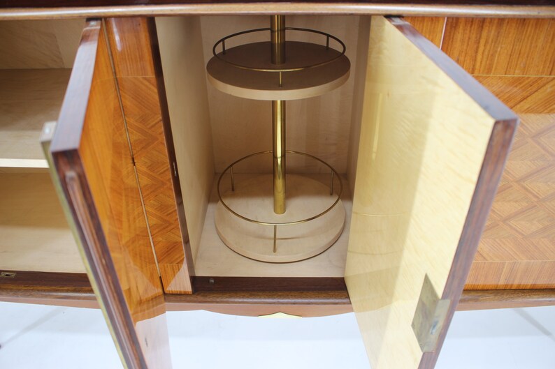 May include: A wooden cabinet with two doors open, revealing a bar area with a rotating shelf and a brass pole. The cabinet has a wood veneer with a geometric pattern.