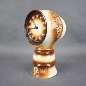 May include: A ceramic table clock with a brown and cream color scheme. The clock face is round with a dark brown border and a cream-colored center. The base is cylindrical with floral patterns.