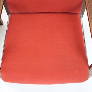May include: Close-up of a mid-century modern chair with a vibrant red seat and backrest. The chair features curved wooden armrests and a sleek, minimalist design. The frame is a dark brown color.
