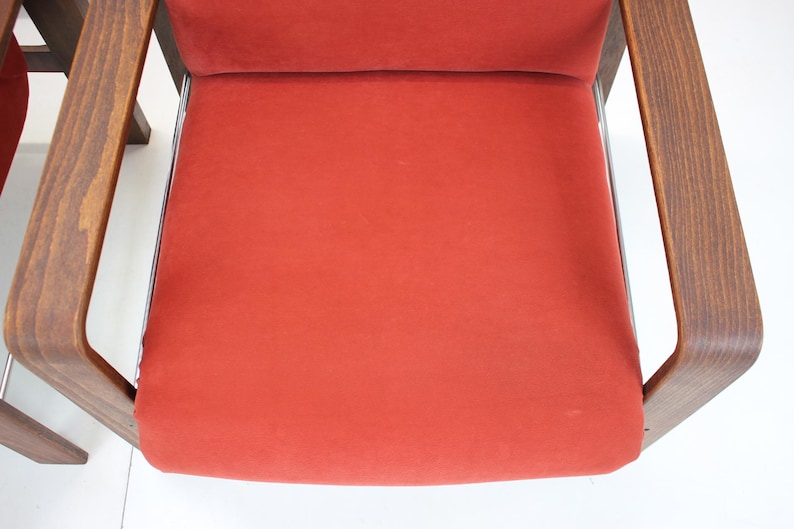 May include: Close-up of a mid-century modern chair with a vibrant red seat and backrest. The chair features curved wooden armrests and a sleek, minimalist design. The frame is a dark brown color.