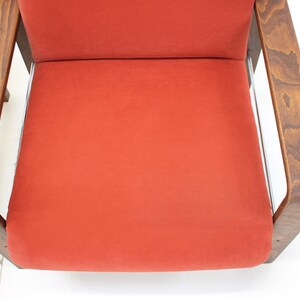 May include: Close-up of a mid-century modern chair with a vibrant orange seat and back. The chair features a wooden frame with a dark finish and curved armrests. The design is simple and elegant, showcasing a retro aesthetic.