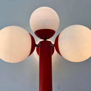 May include: A red and white floor lamp with four spherical white light fixtures. The lamp has a central red pole and red accents around the lights. The background is a light blue.