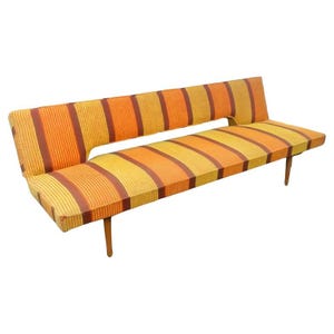 May include: A mid-century modern sofa with a vibrant striped pattern in shades of orange, yellow, and brown. The sofa features a unique asymmetrical design with a low back and wooden legs. The dimensions are approximately 72 inches long.