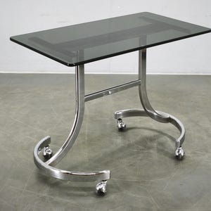May include: A rectangular table featuring a dark grey tinted glass top and a polished chrome metal frame. The base consists of two curved, C-shaped supports connected by a horizontal bar, all mounted on four small swivel castors for mobility.