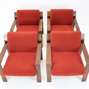 May include: Four mid-century modern armchairs with a warm, reddish-orange seat cushion and dark wood frame. The chairs have a simple, geometric design, with angled armrests and a rectangular back. The chairs are arranged in a symmetrical pattern.