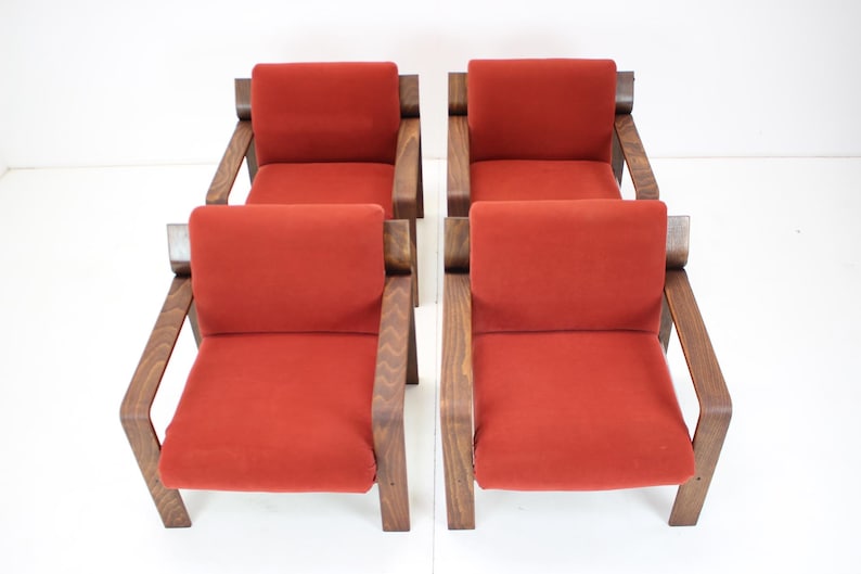 May include: Four mid-century modern armchairs with a warm, reddish-orange seat cushion and dark wood frame. The chairs have a simple, geometric design, with angled armrests and a rectangular back. The chairs are arranged in a symmetrical pattern.