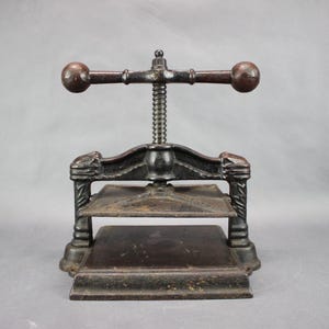 May include: A vintage cast iron book press featuring a dark brown finish, a central threaded screw mechanism, and a horizontal handle with two rounded ends. The device includes a flat base and a movable top plate for applying pressure to books.