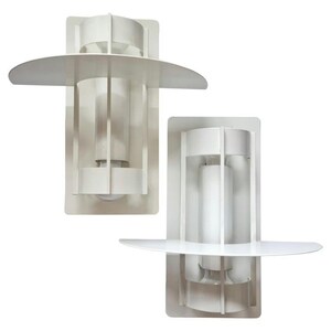 May include: A pair of white wall sconces featuring a rectangular backplate, a central cylindrical light housing, and a horizontal semi-circular shelf extending from the front. The metal structure is composed of vertical slats surrounding the light.