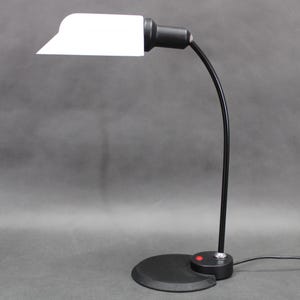 Może przedstawiać: Czarna lampa biurkowa z białym, prostokątnym kloszem. Lampa ma zakrzywione, regulowane ramię i okrągłą podstawę z czerwonym przyciskiem zasilania. Lampa jest podłączona do gniazdka elektrycznego.