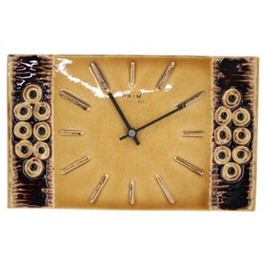 May include: A rectangular ceramic wall clock with a glossy tan face and dark brown textured side panels featuring circular embossed patterns. The clock has black hands and simple raised tan hour markers. The text PRIM CERAMIC is printed in black.