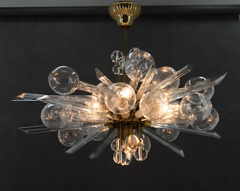 May include: A vintage chandelier with a brass finish and clear glass elements. The fixture features multiple clear glass spheres and pointed glass rods radiating outwards. The design is reminiscent of a burst of light, suitable for interior lighting.