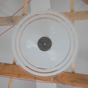 May include: A white frosted glass ceiling light fixture with a circular design and three concentric rings. The light fixture is hanging from a wooden beam.