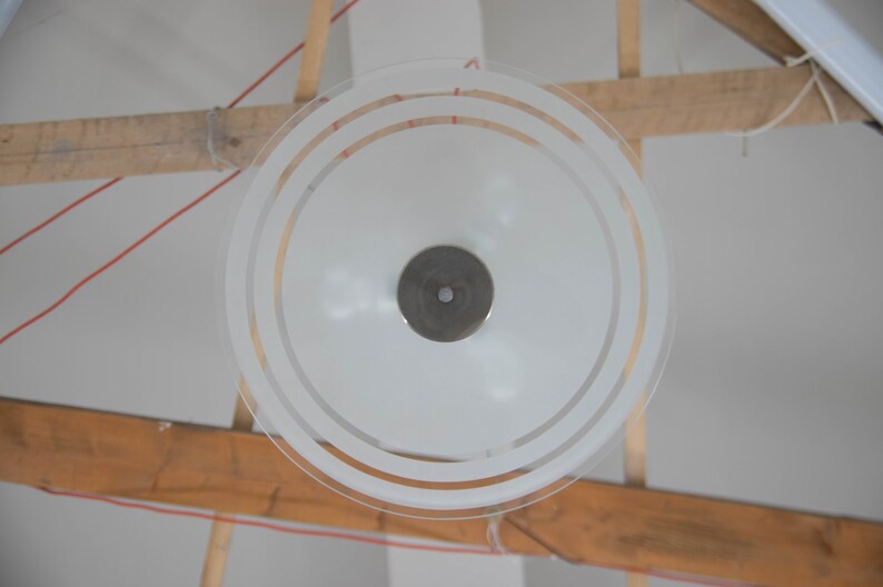 May include: A white frosted glass ceiling light fixture with a circular design and three concentric rings. The light fixture is hanging from a wooden beam.
