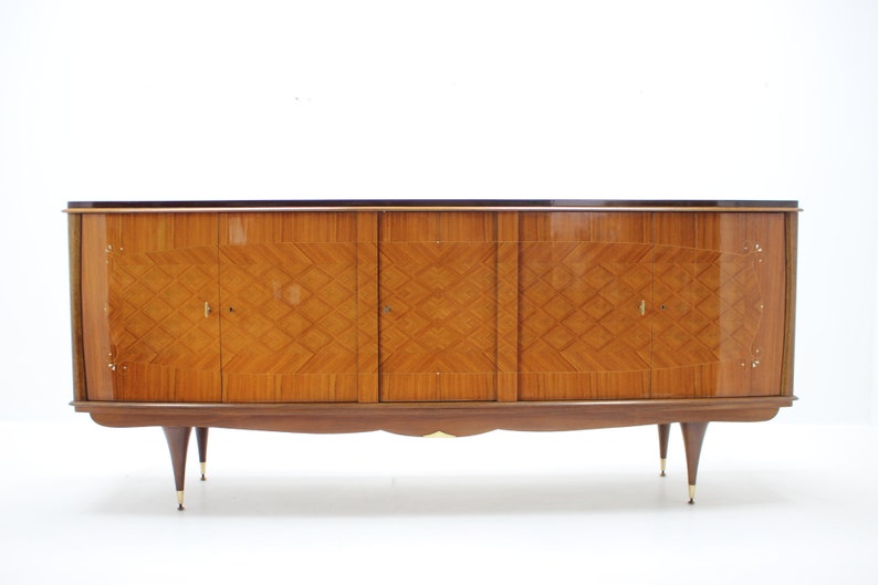 May include: A mid-century modern sideboard with four doors and a wood veneer finish. The cabinet has a dark brown top and sits on four slender legs.