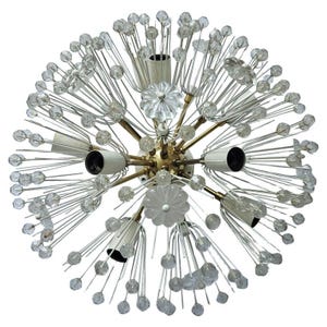 Op de afbeelding: Een kroonluchter in mid-century modern stijl met een sputnik-ontwerp. De lamp heeft een messingkleurige centrale naaf met meerdere armen die naar buiten uitstralen. Elke arm heeft een lamphouder en is versierd met heldere kristallen kralen en bloemvormige accenten.