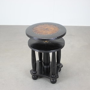 May include: A two-tiered, black wooden side table with a round top and a lower shelf. The top tier features a decorative, inlaid wood design. The table has four curved legs and a decorative base.