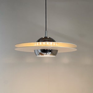 Może przedstawiać: Wisząca lampa chromowana i biała z okrągłym, talerzowym kloszem. Lampa jest zawieszona na czarnym przewodzie.