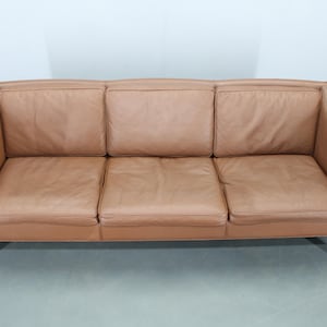 May include: A three-seater sofa upholstered in light brown leather. The sofa features a rectangular design with a gently curved back and armrests. Dark wood legs support the sofa.
