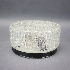 May include: A round flush mount ceiling light fixture featuring a clear, textured glass shade with a bubbly, organic pattern. The glass shade is secured to a solid black circular metal base, creating a contrast between the clear top and dark bottom.