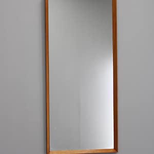 May include: Rectangular mirror with a light brown wooden frame. The mirror is mounted on a gray wall. The frame is a simple, clean design, and the mirror itself is clear and reflective.