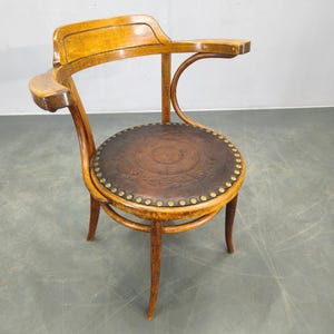 May include: A vintage wooden armchair with a curved back and armrests. The round seat is made of brown leather and is adorned with decorative brass studs. The chair has a warm, natural wood finish.