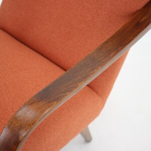 Pode incluir: Um close-up de uma poltrona laranja com bra&ccedil;os de madeira. A cadeira tem um encosto curvo e um design simples.