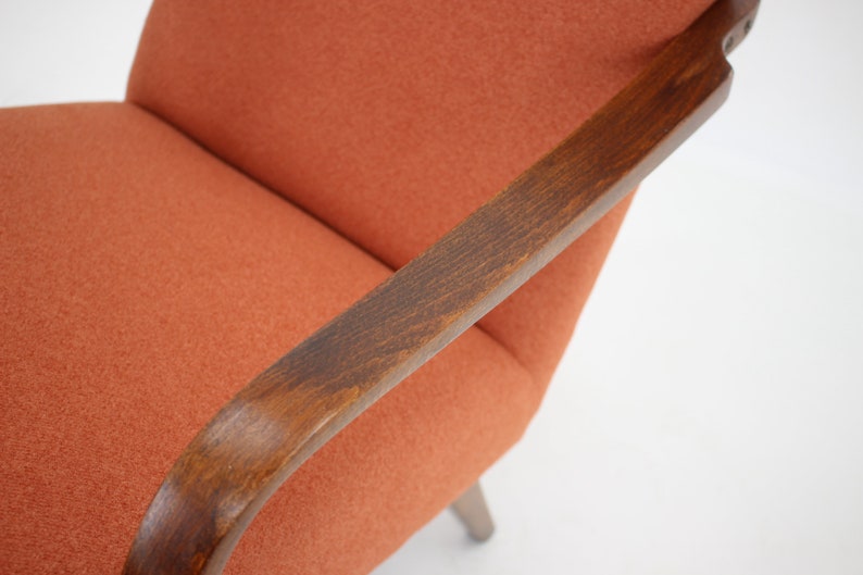 Pode incluir: Um close-up de uma poltrona laranja com bra&ccedil;os de madeira. A cadeira tem um encosto curvo e um design simples.