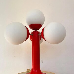 May include: A retro-style table lamp with a vibrant red base and stem, featuring four spherical white glass shades. The lamp's design is reminiscent of mid-century modern aesthetics, adding a pop of color and unique lighting to any space.