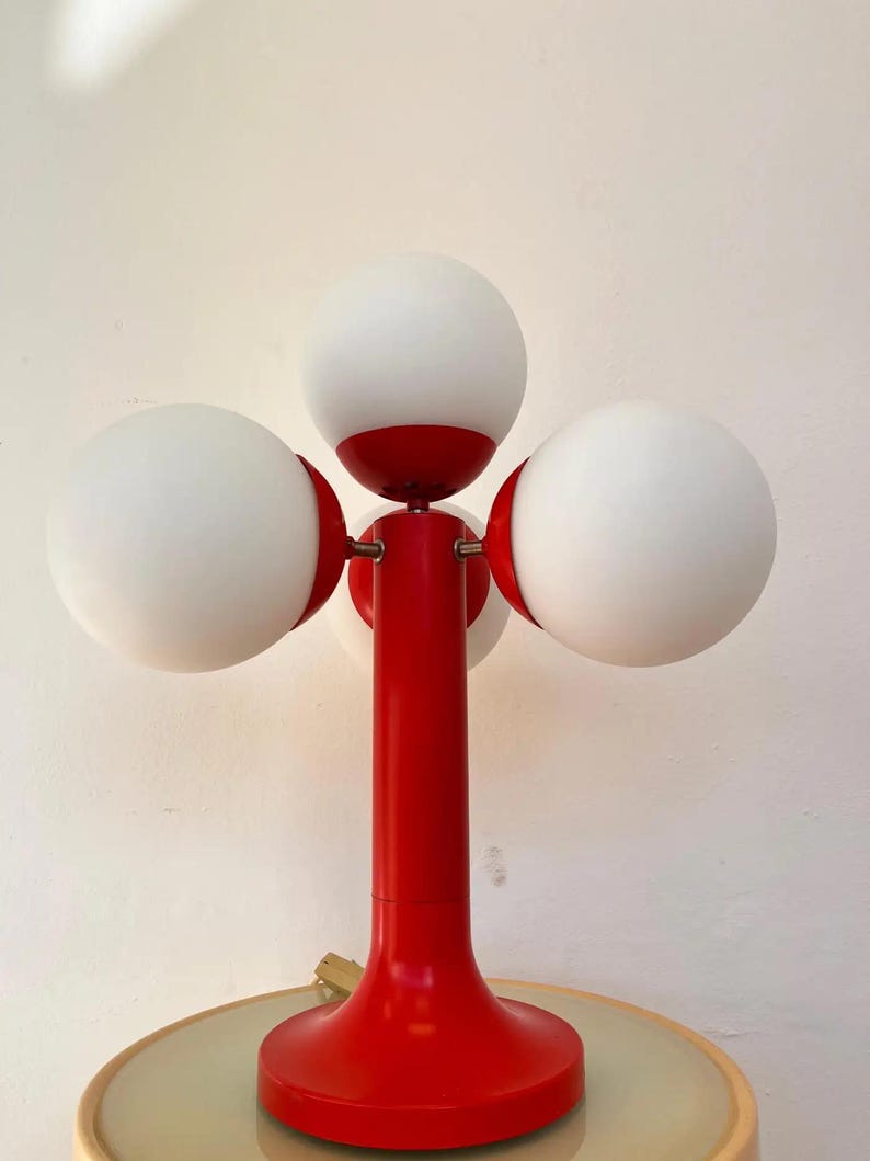 May include: A retro-style table lamp with a vibrant red base and stem, featuring four spherical white glass shades. The lamp's design is reminiscent of mid-century modern aesthetics, adding a pop of color and unique lighting to any space.