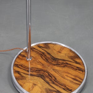 May include: A vintage floor lamp base with a circular wooden base featuring a striking wood grain pattern. The base is encased in a silver metal rim, and a chrome pole extends upwards. The lamp's design is mid-century modern.