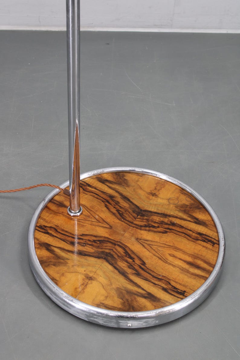 May include: A vintage floor lamp base with a circular wooden base featuring a striking wood grain pattern. The base is encased in a silver metal rim, and a chrome pole extends upwards. The lamp's design is mid-century modern.