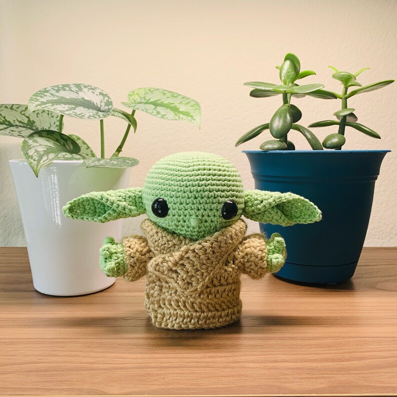 May include: A crocheted green and brown Yoda doll with large eyes and a brown robe. The doll is sitting on a wooden surface in front of two potted plants.