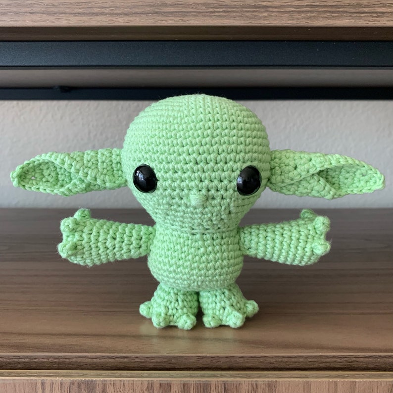 May include: A green crocheted plush toy resembling a popular fictional character. The toy has large, black eyes and a small, pointed nose.