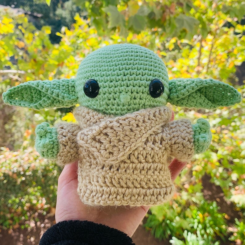 May include: A crocheted green and brown plush toy resembling a character from the Star Wars franchise. The toy has large eyes and a small mouth, and is wearing a brown crocheted sweater.