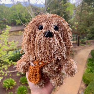 May include: A brown and white plush toy dog with a brown nose and black eyes. The dog is wearing a brown and orange crocheted vest.