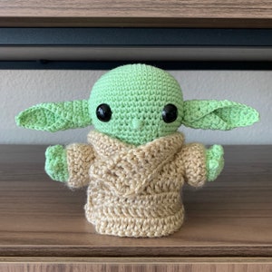 May include: A crocheted green and brown plush toy resembling a character from the Star Wars franchise. The toy has large black eyes and a small mouth.