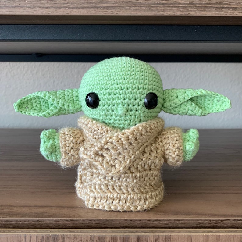 May include: A crocheted green and brown plush toy resembling a character from the Star Wars franchise. The toy has large black eyes and a small mouth.